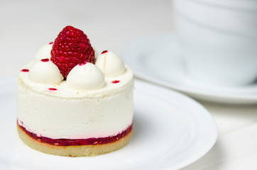 White airy cake with raspberry layer and raspberry berry lies on a white round plate next to a white cup, which stands on a white table