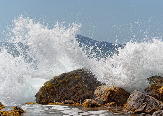 A wave breaks on big stones in the water, thousands of drops of water sparkle in the sunlight, island silhouette in the background, snapshot - Location: Caribbean, island Guadeloupe