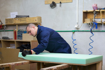 Young man in a furniture factory applies a foam on a wooden piece of the sofa