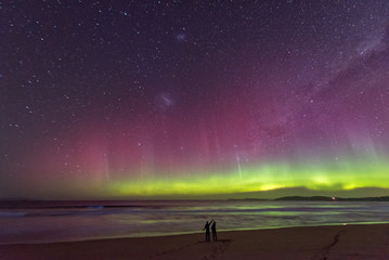 Two people in silhouette standing in awe of an incredible display of the Aurora Australis or Southern Lights, with bioluminescence turning the breaking waves bright blue.