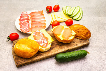 Homemade sandwich in bun, step by step preparation. Salami, dried pork, tomato, cucumber, cheese ingredients. On wooden board and stone background.
