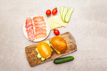 Homemade sandwich in bun, step by step preparation. Salami, dried pork, tomato, cucumber, cheese ingredients. On wooden board and stone background, top view.