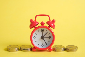 The concept of saving money in a crisis. A red vintage clock stands on a stack of coins on a yellow background.