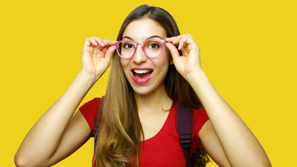 Portrait of a happy casual girl student with backpack holding her glasses standing over yellow background