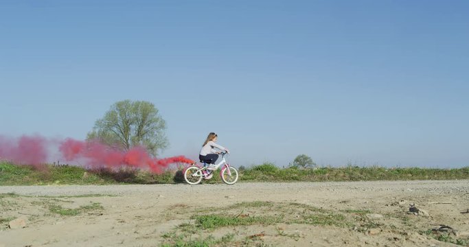 Authentic shot of cute little girl carefree having fun and going with her bicycle with a colorful smoke flare on a countryside background in a sunny day.