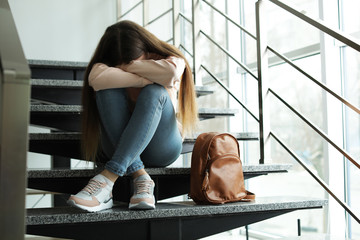 Upset teenage girl with backpack sitting on stairs indoors