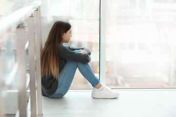Upset teenage girl sitting at window indoors. Space for text