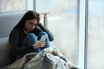 Upset teenage girl with smartphone sitting at window indoors. Space for text