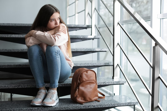 Upset teenage girl with backpack sitting on stairs indoors. Space for text - Powered by Adobe