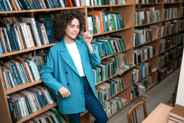 Young beautiful curly girl in glasses and a blue suit is standing in the library. Student Study