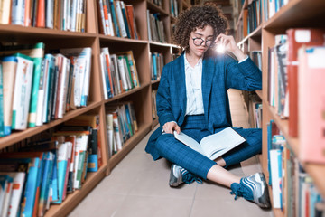 Young beautiful curly girl in glasses and a blue suit is standing in the library. Student Study