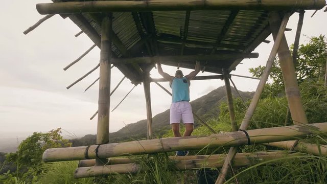Handsome man training pull up exercise on wooden crossbar outdoor. Athlete man exercising pull ups on green hill landscape. Workout gym.
