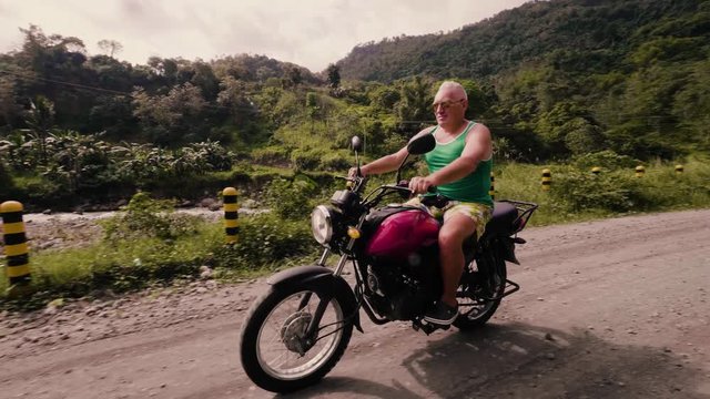 Senior man riding on motorcycle on road while traveling at summer vacation. Elderly motorcyclist driving on motorbike on green hills and highlands covered tropical forest.