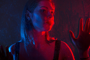 Glamorous brown-haired woman outside the window with drops of water in neon light close-up.