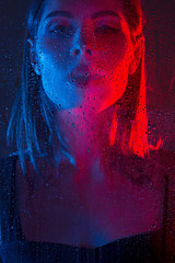 Glamorous brown-haired woman breathes on a window with water drops in neon light close-up.