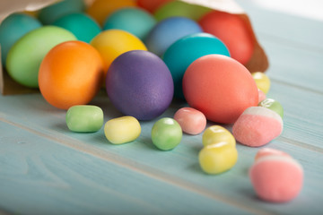 Easter colorful dyed chicken eggs in a paper bag and sweets on a blue wooden table.