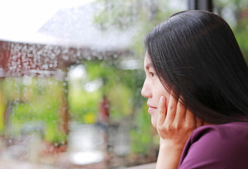 Beautiful Asian woman looking out from the glass window and feeling happy during rainy day.