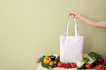 Female hand with eco bag and fresh vegetables on table against color background