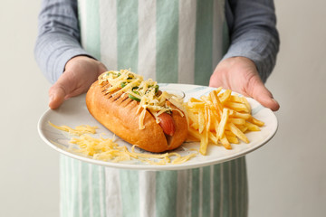 Woman holding plate with hot dog and french fries on light background