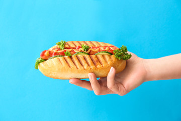 Female hand holding tasty hot dog on color background