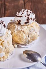 Two airy desserts with meringue in chocolate drops with prunes and butter cream in a white plate next to tea spoon and napkin on a wooden background. Close-up
