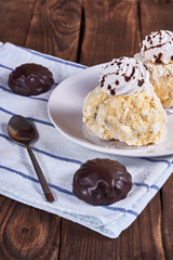Two airy desserts with meringue in chocolate drops with prunes and butter cream and the chocolate zephyr in a white plate next to tea spoon and napkin on a wooden background. Close-up