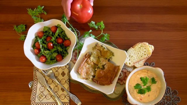 Healthy mediterranean cuisine, three dishes concept for lunch or dinner. Closeup of placing a tomato next to the letuce salad on the table in slow motion