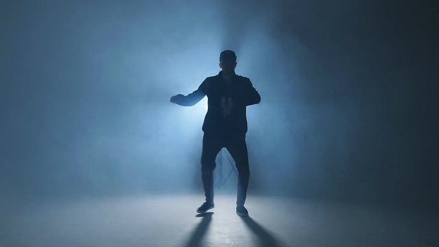 A dark silhouette of a singer on the stage, dancing alone during performance on dark blue neon background with smoke and lights, slow motion.