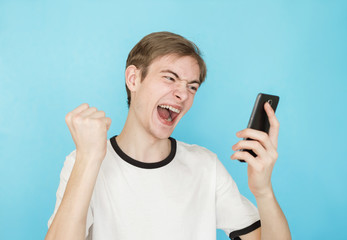 Fototapeta premium young funny male teenager in white t-shirt on blue background looks into smartphone as if won something