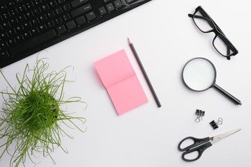 White table with keyboard, glasses, magnifying glass, pink stickers, clips, scissors, office plant and pencil.