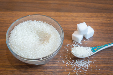 White sugar kept in a glass bowl with a spoon. Some sugar cubes kept on a clean wooden floor. Sugar taken out from a bowl with a spoon.