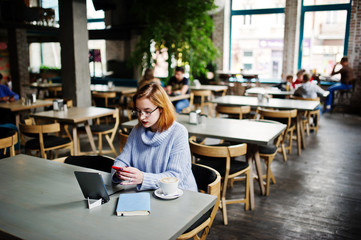 Cheerful young beautiful redhaired woman in glasses using her phone, touchpad and notebook while sitting at her working place on cafe with cup of coffee.
