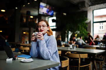 Cheerful young beautiful redhaired woman in glasses using her phone, touchpad and notebook while sitting at her working place on cafe with cup of coffee.