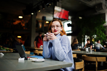 Cheerful young beautiful redhaired woman in glasses using her phone, touchpad and notebook while sitting at her working place on cafe with cup of coffee.