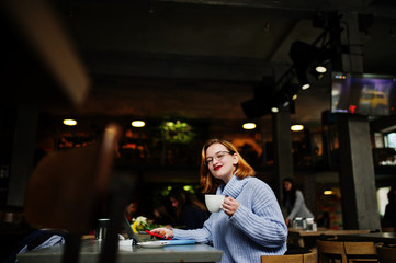 Cheerful young beautiful redhaired woman in glasses using her phone, touchpad and notebook while sitting at her working place on cafe with cup of coffee.