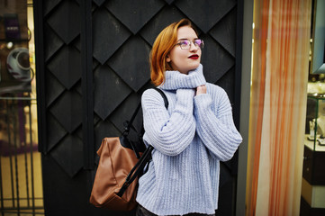 Cheerful young beautiful redhaired woman in glasses, warm blue wool sweater with backpack posed outdoor.