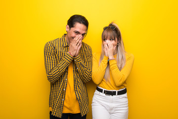 Young couple over vibrant yellow background smiling a lot while covering mouth