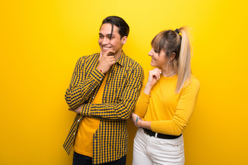 Young couple over vibrant yellow background looking to the side with the hand on the chin