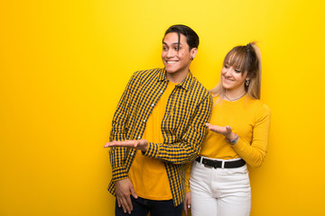 Young couple over vibrant yellow background presenting an idea while looking smiling towards
