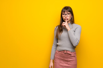 Woman with glasses over yellow wall showing a sign of silence gesture putting finger in mouth