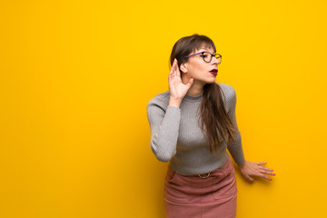 Woman with glasses over yellow wall listening to something by putting hand on the ear