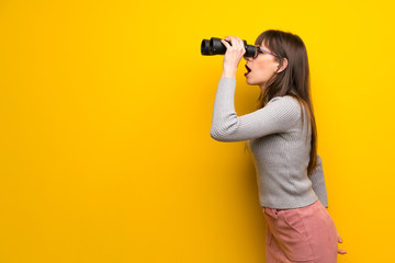 Woman with glasses over yellow wall and looking in the distance with binoculars