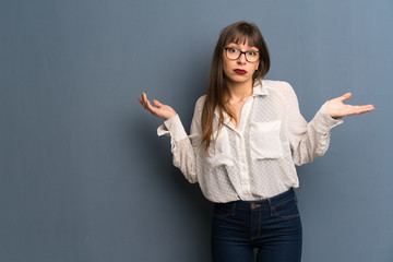 Woman with glasses over blue wall having doubts while raising hands and shoulders
