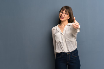 Woman with glasses over blue wall giving a thumbs up gesture because something good has happened