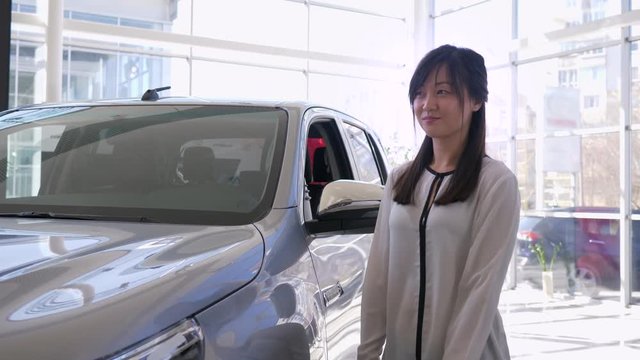 Auto business, portrait of smiling saleswoman pats car with pleasure and shows keys to new automobile for sale in sales center close-up