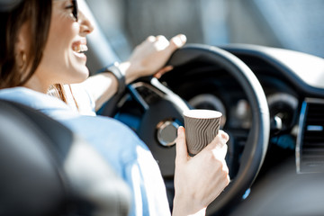 Cheerful woman holding steering wheel and coffe cup while driving a car, close-up view