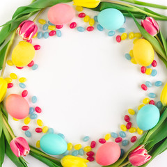 Colorful Easter wreath made of yellow tulips, colorful eggs and candies on white background. Flat lay.