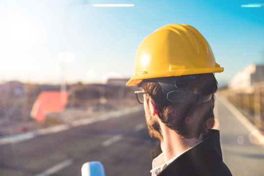 Ingegnere Edile in un cantiere osserva i lavori e il progetto per assicurarsi che vada tutto bene. Visto di spalle con caschetto giallo.