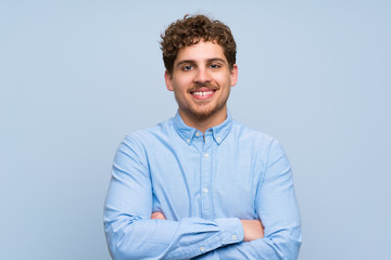 Blonde man over blue wall keeping the arms crossed in frontal position