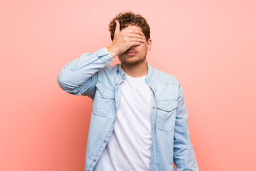 Blonde man over pink wall covering eyes by hands. Do not want to see something
